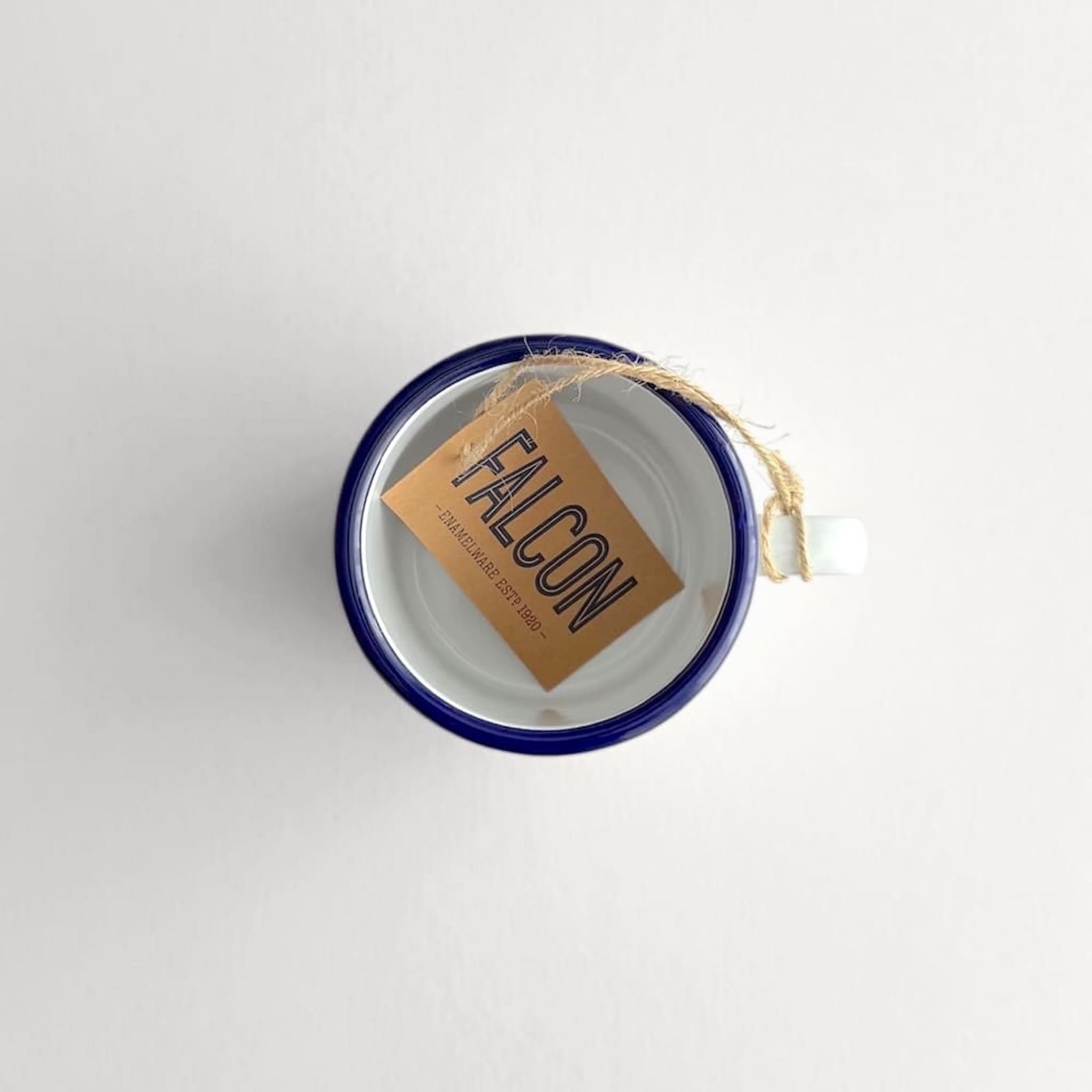 Above view of a white Falcon enamelware mug with blue rim and kraft FALCON logo tag