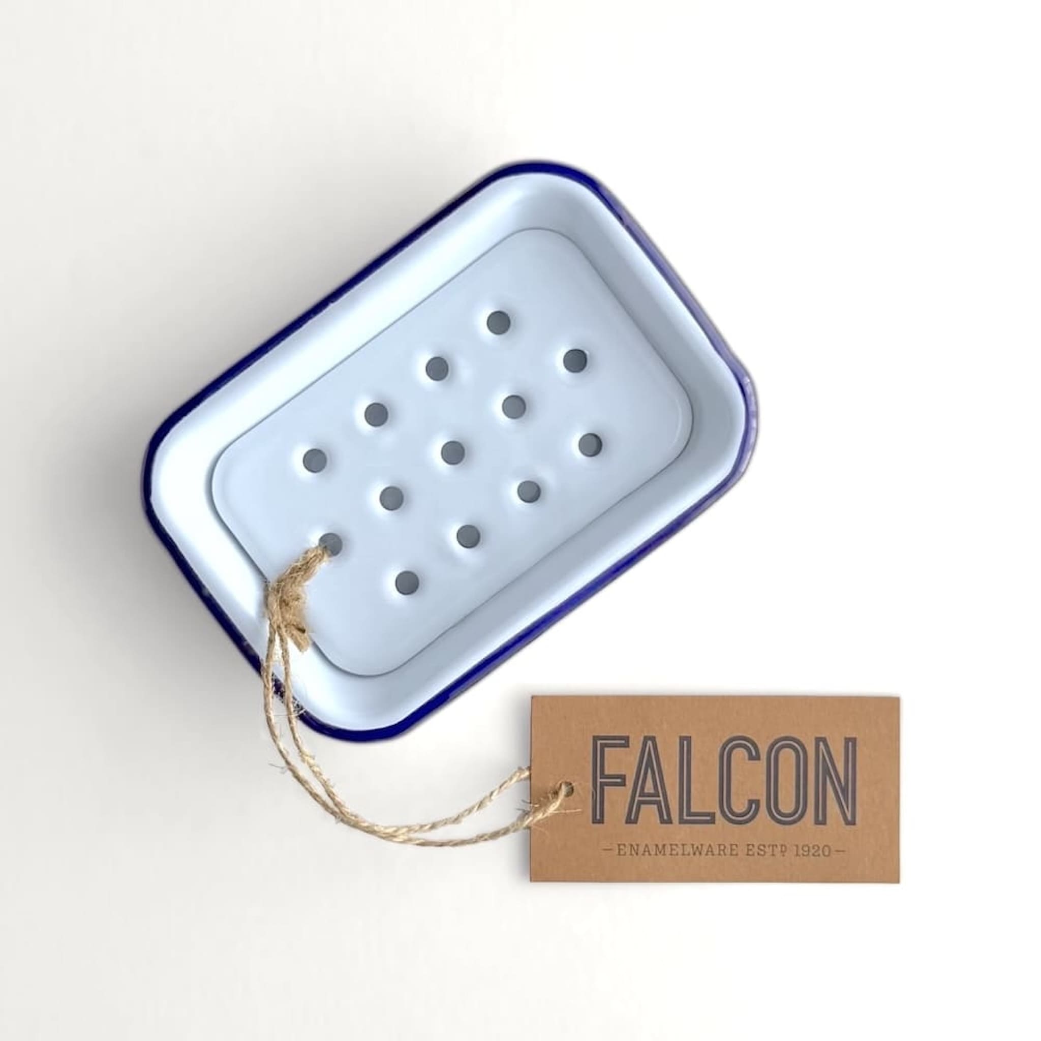 White enamel soap dish with blue rim and hanging tag on a white background, featuring the brand 'Falcon'