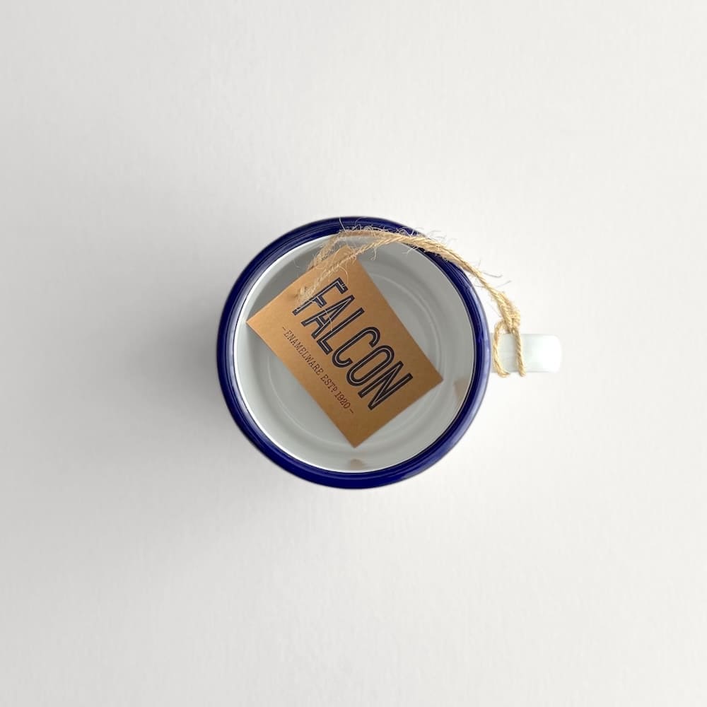 Above view of a white Falcon enamelware mug with blue rim and kraft FALCON logo tag 