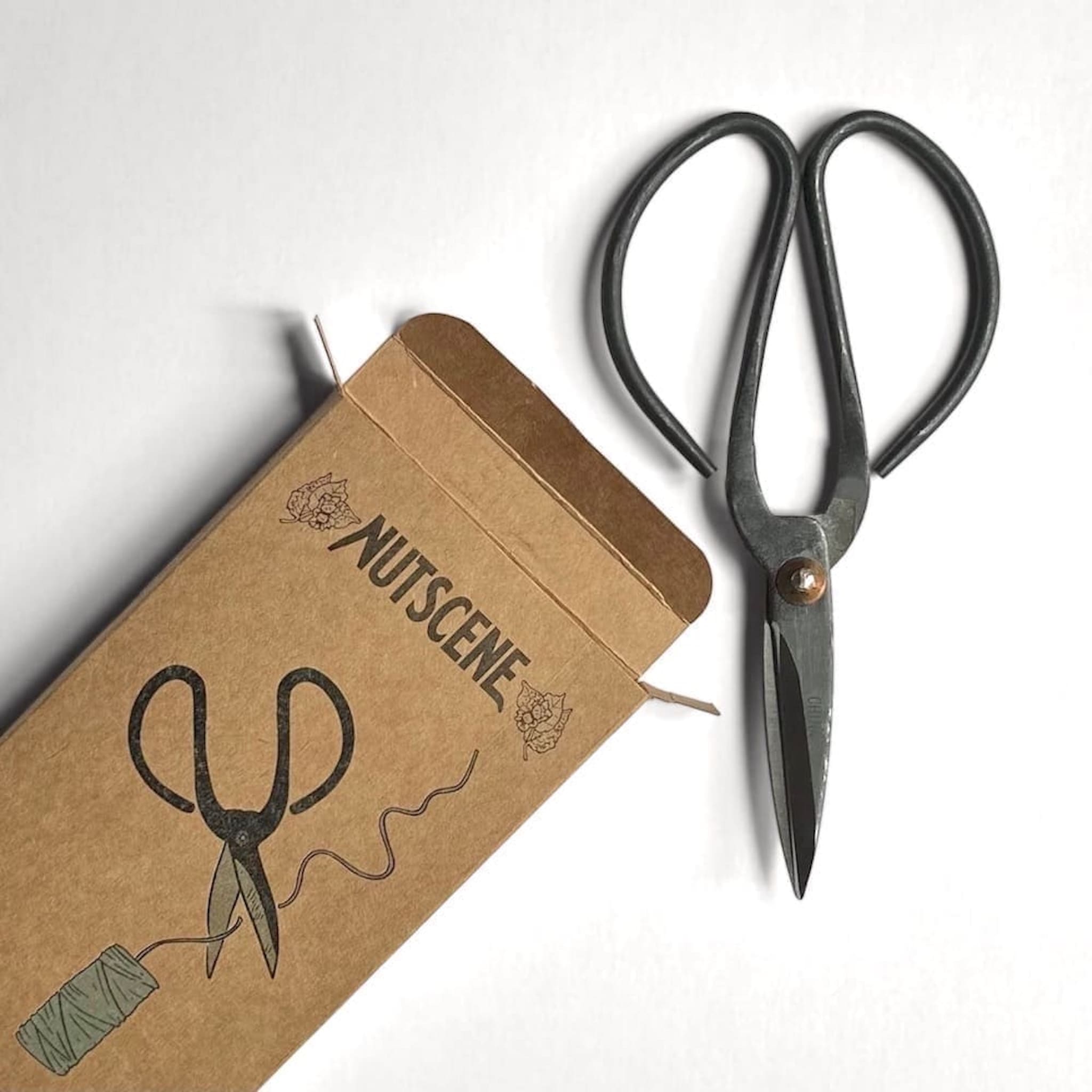 Pair of black-handled scissors next to a brown box with 'NUTSCENE' branding on a white background