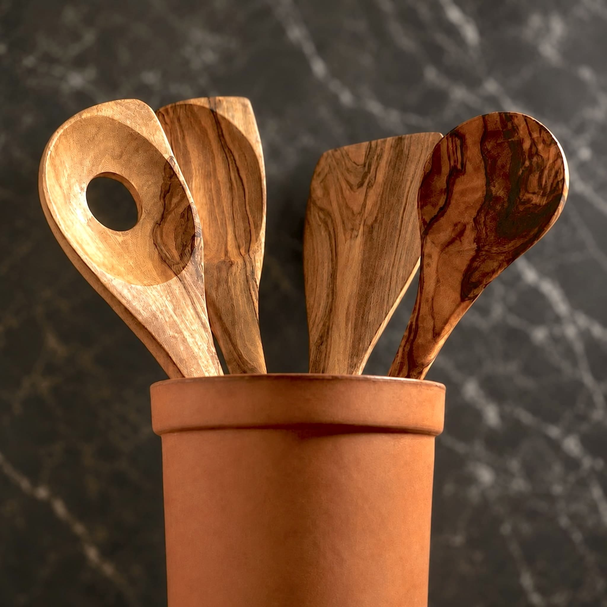 Four wooden kitchen utensils with a rich grain pattern, resting in a terracotta holder against a dark marble background.