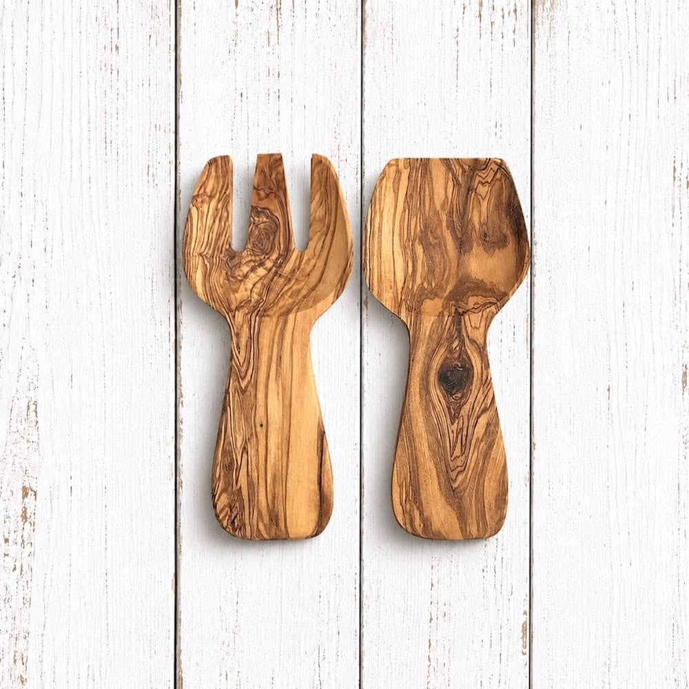 Two olive wood salad serving utensils with natural grain patterns, displayed side-by-side on a white distressed wood plank background.