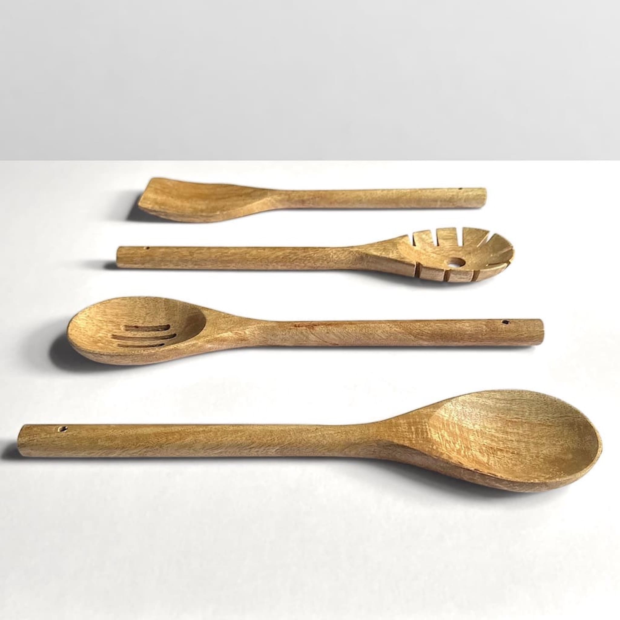 Set of four mango wood kitchen utensils including a spoon, slotted spoon, pasta spoon and and spatula on an off-white background